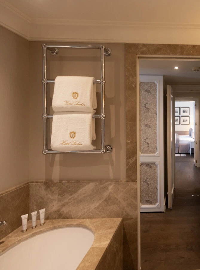 A warmly lit bathroom at Hotel Sacher Salzburg features a marble bathtub, a chrome towel warmer with folded white towels, a double sink vanity with marble countertops, and a glimpse into an adjacent room with a bed.