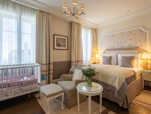 A warmly lit hotel room at Hotel Sacher Salzburg features a double bed, crib, armchair, and city view through the window.