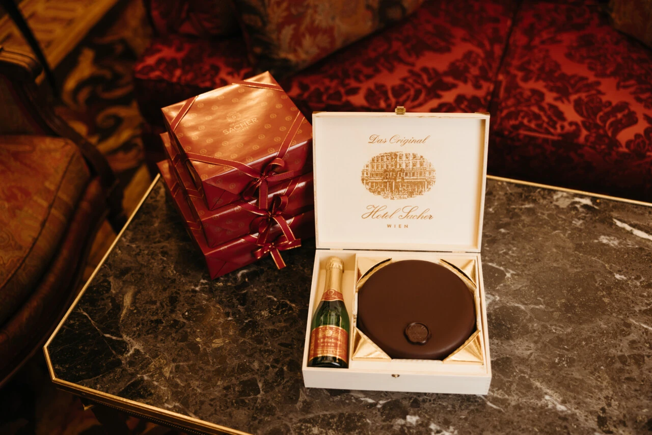 A Sacher Torte in its box, next to a bottle of Sacher Cuvée and some gifts, likely from Hotel Sacher in Vienna.