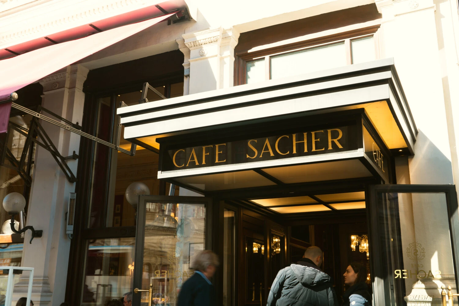 Cafe Sacher Wien entrance: Illuminated sign above the doorway of the famous Viennese cafe, with people entering and exiting.