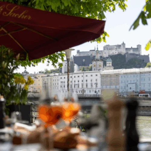 View of Hohensalzburg Fortress from a riverside restaurant table with Aperol Spritz and sunlit dining setup
