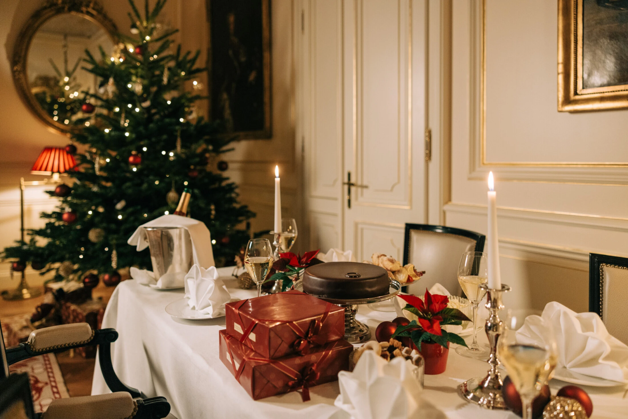 A festive Christmas dinner table with wrapped gifts, candles, and poinsettias, set against a backdrop of a decorated Christmas tree and elegant room.