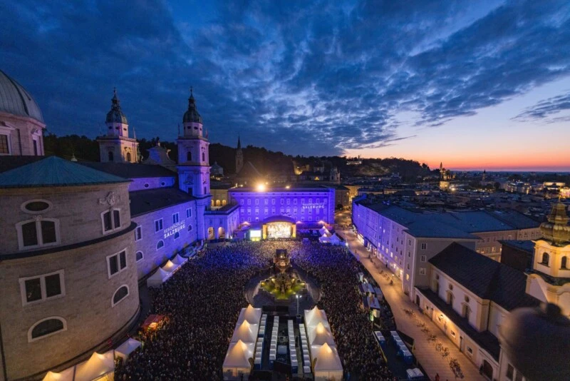 pizzera-&-jaus-concert-2026-salzburg Aerial view of Salzburg's Cathedral Square at dusk, filled with a crowd and illuminated by blue and purple lights.