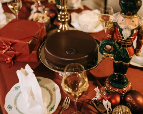 festive table set with a red tablecloth, featuring a glossy chocolate cake on a silver stand in the center. To the right, part of a nutcracker figure and red Christmas ornaments are visible.