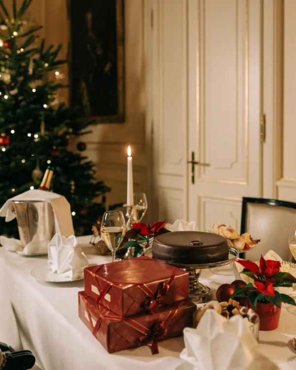 A festive Christmas dinner table with wrapped gifts, candles, and poinsettias, set against a backdrop of a decorated Christmas tree and elegant room.
