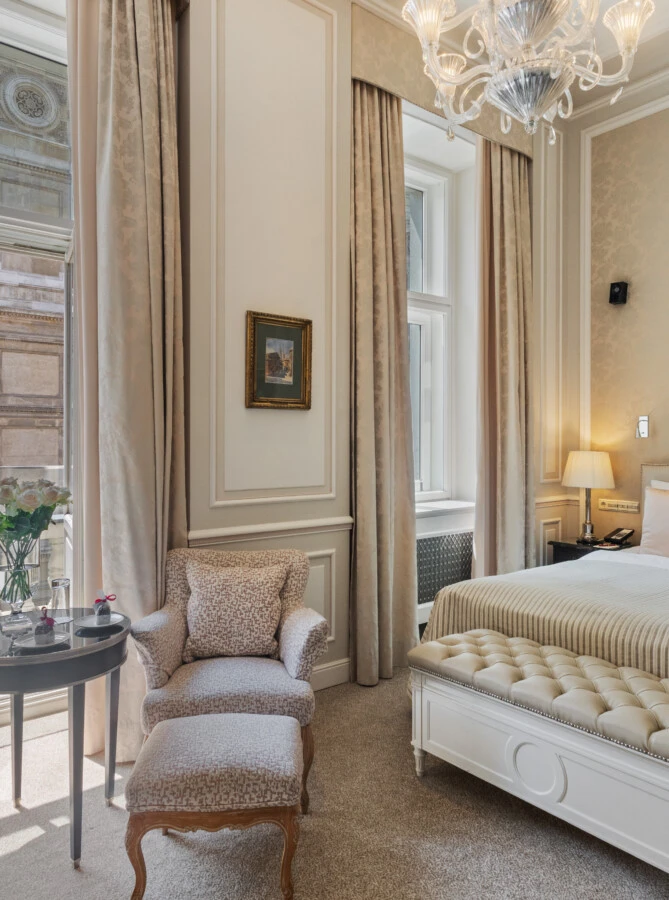 A beautiful hotel room featuring a bed, elegant furnishings, and a stunning balcony view of the Vienna State Opera (Wiener Staatsoper).