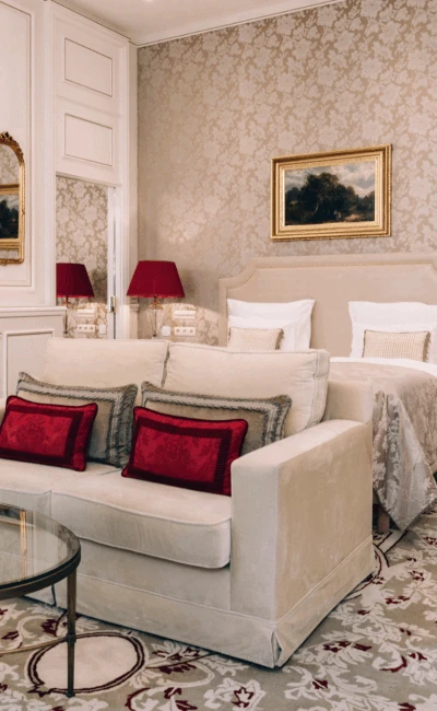 A beautifully decorated junior suite with a combined sleeping and living area, featuring a sofa with red cushions and a classic bed.