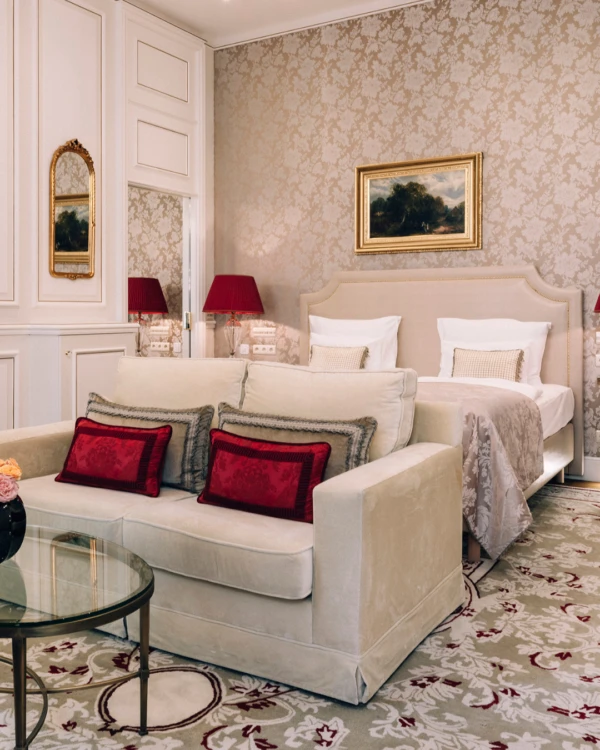 A beautifully decorated junior suite with a combined sleeping and living area, featuring a sofa with red cushions and a classic bed.