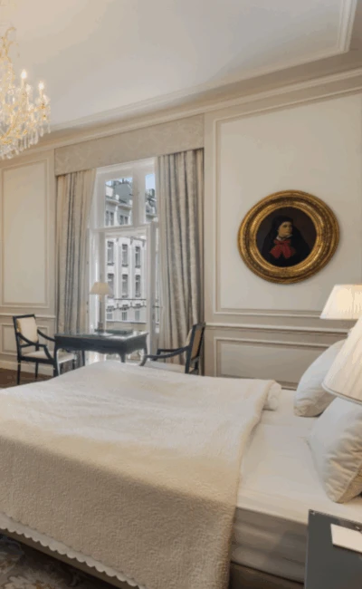 A luxurious and spacious hotel room with a bed, a sofa seating area, a writing desk, and a fireplace under a crystal chandelier.