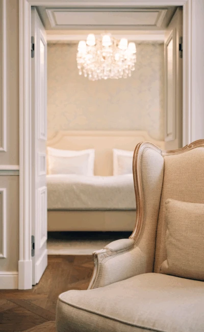 A view from an elegant sitting room into a luxurious bedroom suite, with a plush armchair in the foreground and a chandelier and bed visible through an open doorway