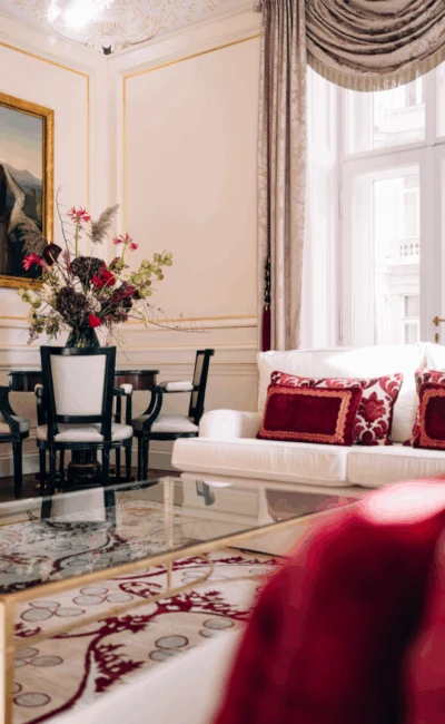A luxurious hotel suite living room with a white sofa, red pillows, a glass coffee table, and a large painting on the wall.
