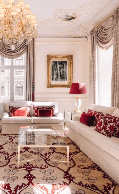 A luxurious hotel suite living room with two white sofas, a patterned rug, and portraits on the walls.