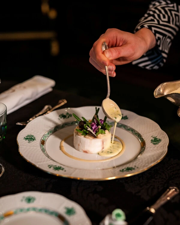A waiter pours a creamy sauce over a fish dish garnished with flowers and herbs on a white and green plate.