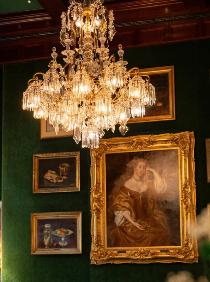 A golden crystal chandelier hangs in front of a green wall decorated with various framed paintings, including a large portrait of a seated woman.