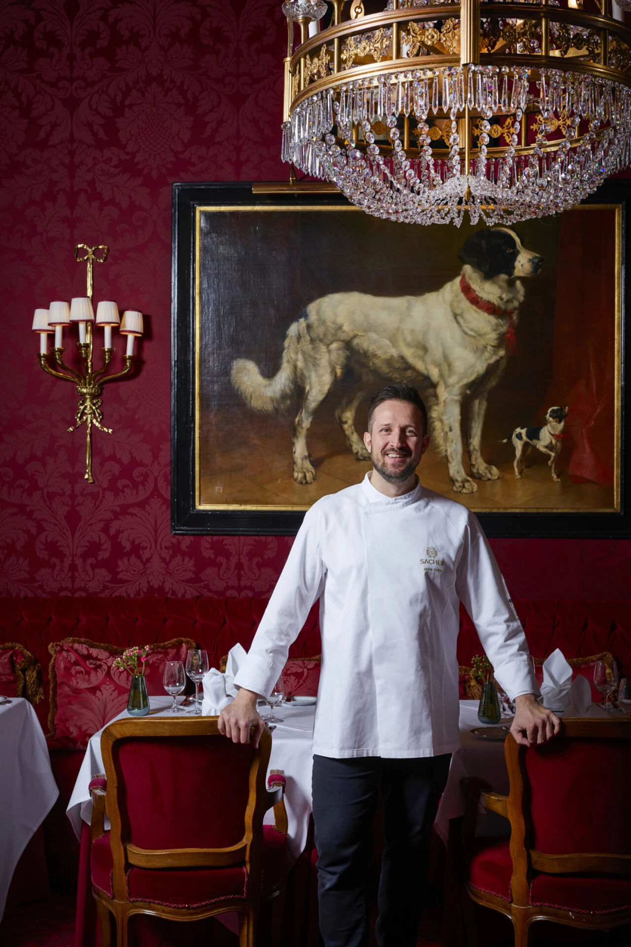 Head chef at Hotel Sacher Vienna standing in the elegant restaurant with red velvet chairs, set tables, and a historic painting in the background