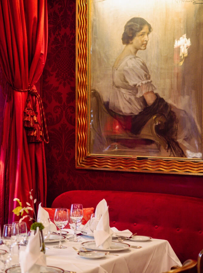 Elegantly set table at Hotel Sacher Vienna with white cloth napkins, crystal glasses and porcelain, in front of red velvet curtains and a historic portrait painting.