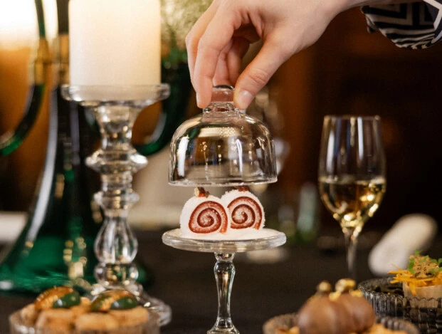 Hand lifts glass dome over small red sponge roll on a festively set table.