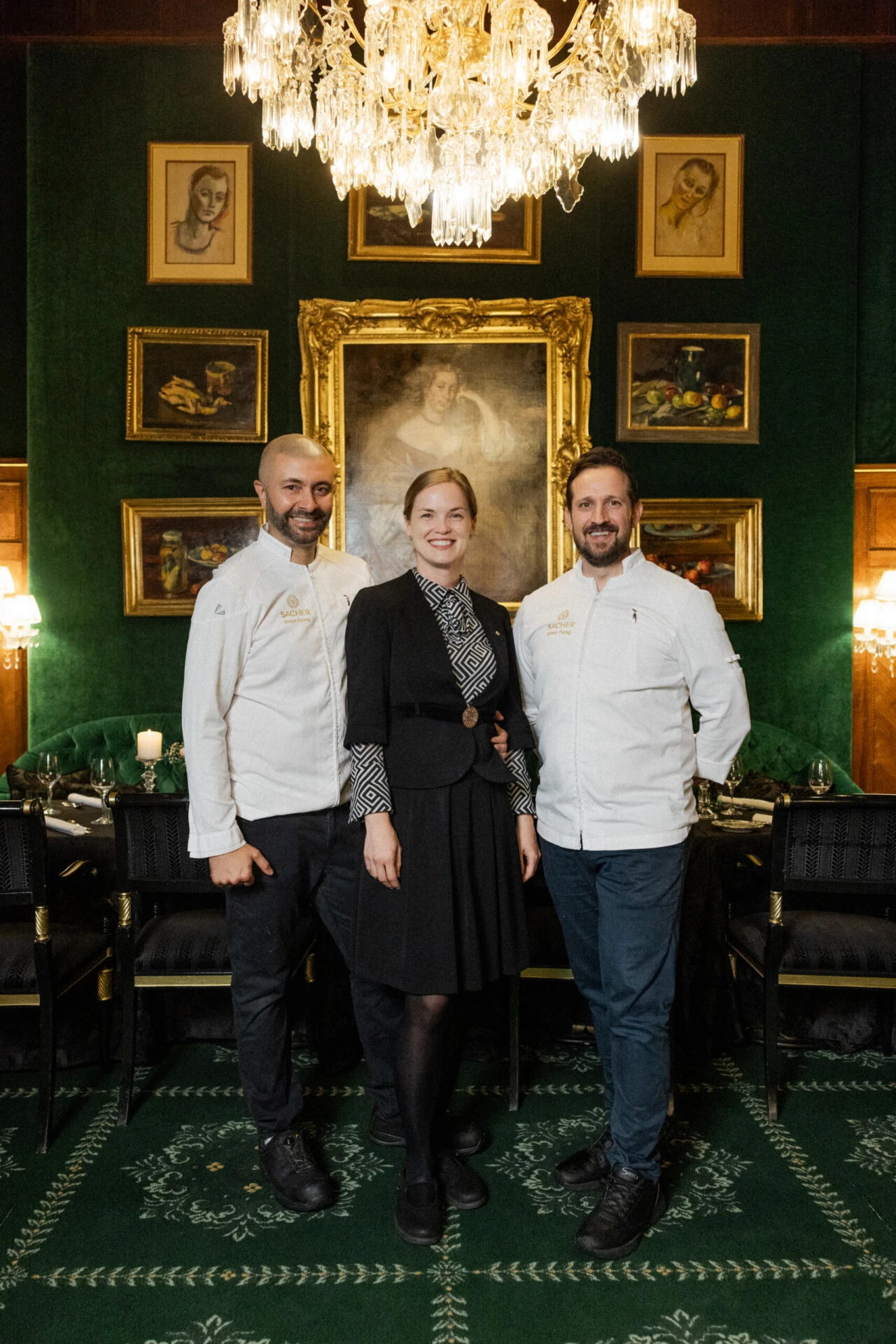 Three people smiling in an elegant room with chandelier and paintings.