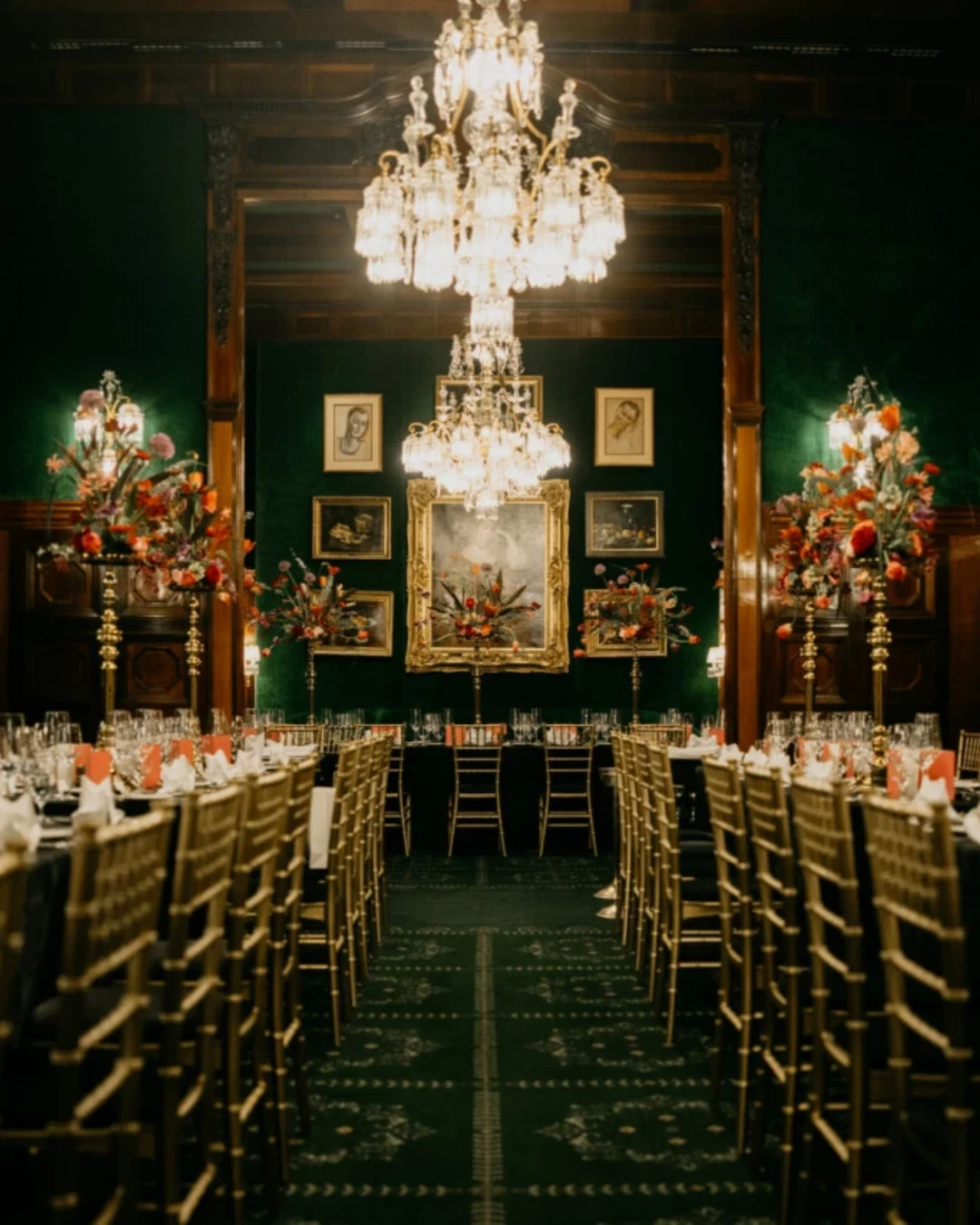 Long table set for a formal gala dinner in a luxurious room with dark green velvet walls, chandeliers, and high floral arrangements.
