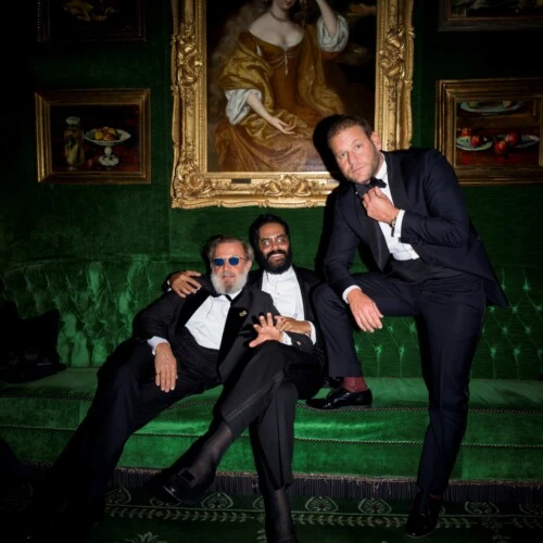 A group of three men in black tie attire are seated and standing on a deep green velvet sofa beneath a large, ornate portrait in a lavish, art-filled room.