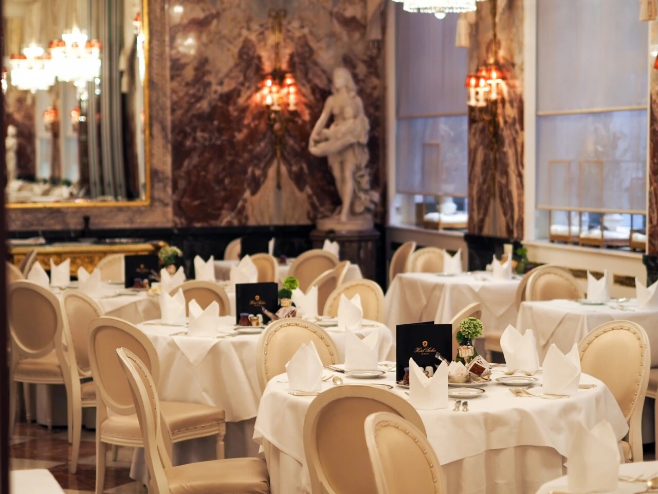 Elegant restaurant with white tablecloths, cream-colored upholstered chairs, and gold accents; marble columns, wall mirrors, chandeliers, and a classic sculpture in the background.