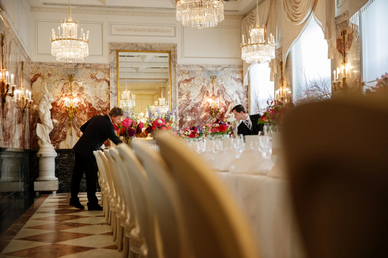 A large, magnificent ballroom with crystal chandeliers, marble columns, and ornate wall decorations, where staff prepare elegant tables with white tablecloths and colorful flower arrangements.