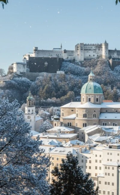 Snow-covered Salzburg Old Town with Hohensalzburg Fortress on the hill, framed by snowy branches.