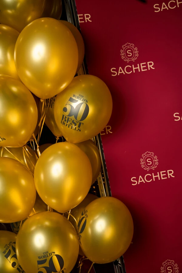 A cluster of gold balloons against a deep red background with the SACHER logo repeated in gold. Some balloons feature the black logo for 