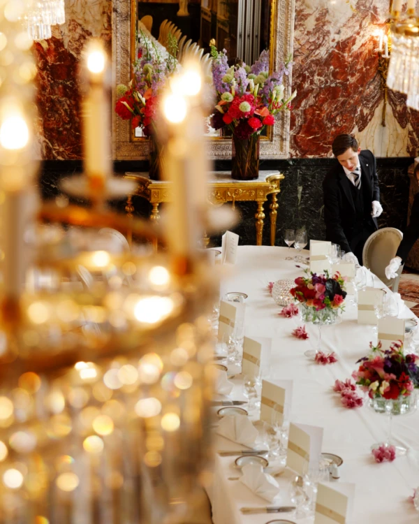 Under glittering chandeliers and rich marble walls, two staff members in formal attire carefully prepare an opulent dining table adorned with fresh flowers and elegant place settings at Hotel Sacher Wien.