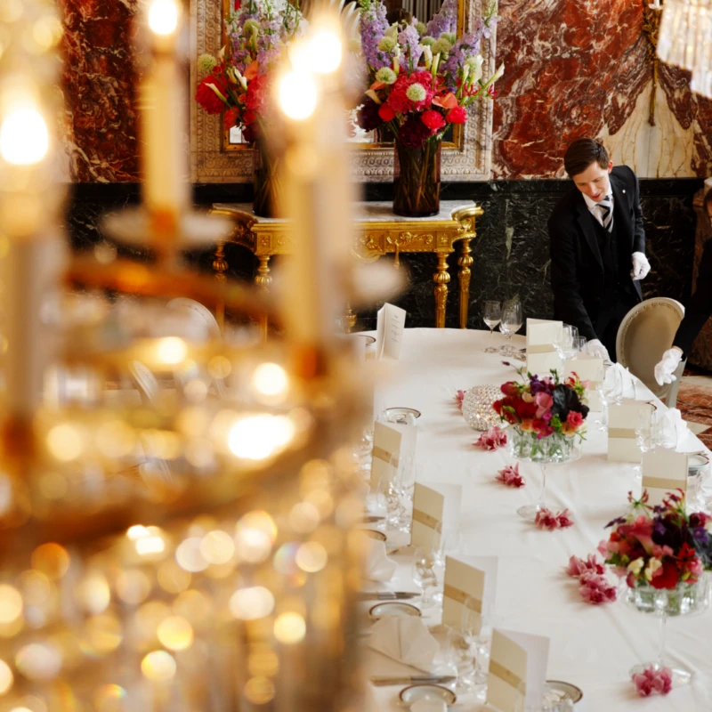 Under glittering chandeliers and rich marble walls, two staff members in formal attire carefully prepare an opulent dining table adorned with fresh flowers and elegant place settings at Hotel Sacher Wien.