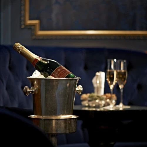 Silver champagne cooler with a bottle of Louis Roederer Cristal Brut on ice in front of a dark blue quilted velvet bench. In the background, on a round table, two filled champagne glasses, a napkin, and a bowl of snacks.