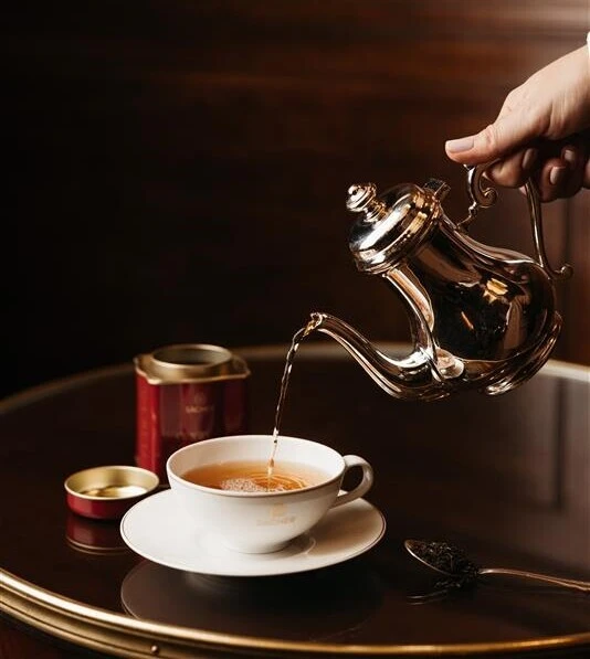 Elegant photo of a silver teapot pouring tea into a white Sacher branded teacup next to a red Sacher tea tin on a polished wooden table.