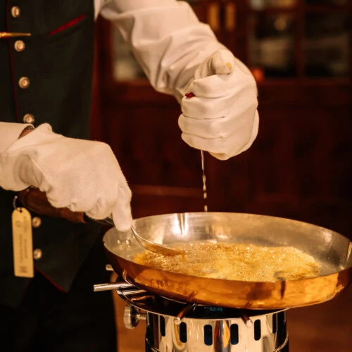 Close-up of a white-gloved server squeezing lemon juice over a thin crepe cooking in a copper pan on a tableside flambé burner.