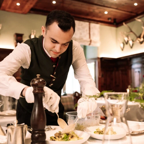 Focused waiter in Austrian formal vest and white gloves preparing a fresh salad or side dish tableside in a luxury restaurant.