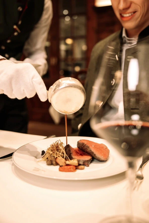 Close-up of a white-gloved server pouring rich jus or sauce over a perfectly cooked beef or venison roast at a fine dining table.