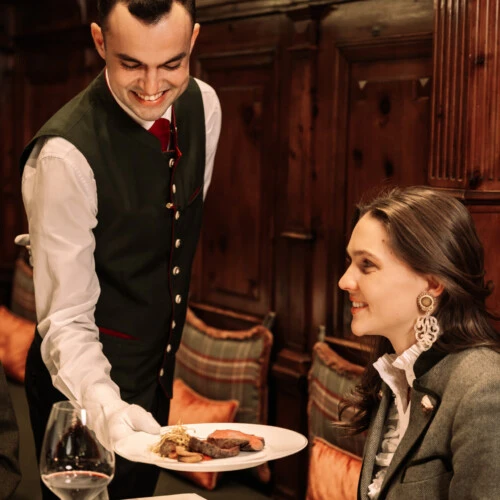 Smiling waiter in traditional Austrian formal vest serving an elegant female guest at a table in a wood-paneled, luxury fine-dining room.