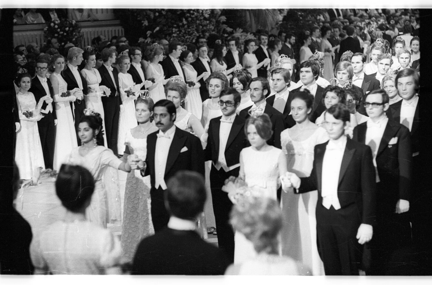 Black and white photo of debutantes and escorts in formal white tie and ball gowns, lining a grand ballroom floor at a historical ball or formal reception.