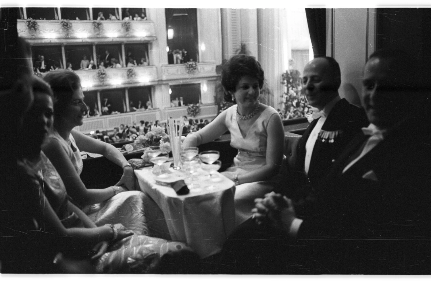 Historical black and white photo of guests socializing and having drinks at a small table, overlooking a crowded, ornate ball or opera house from a balcony or loge.