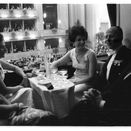 Historical black and white photo of guests socializing and having drinks at a small table, overlooking a crowded, ornate ball or opera house from a balcony or loge.