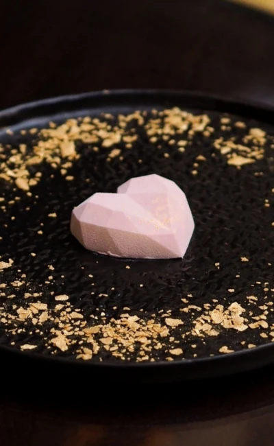 Heart-shaped, pink praline with a geometric pattern, presented on a black plate with shiny gold sprinkles.