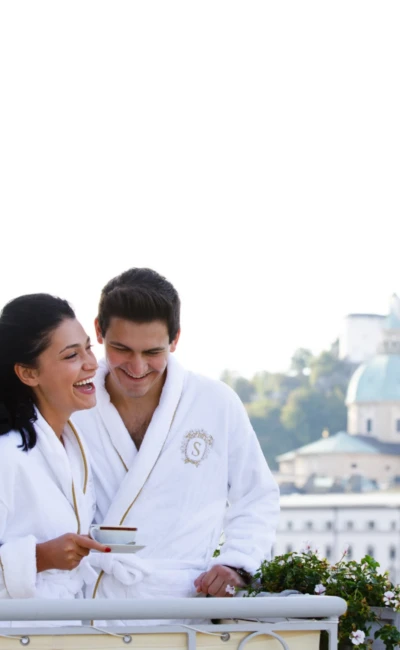 A couple in white bathrobes with the Sacher logo are standing on the balcony. They are both laughing and enjoying a cup of coffee together. In the background, you can see the silhouette of Salzburg with the Hohensalzburg Fortress and the domes of the cathedral.