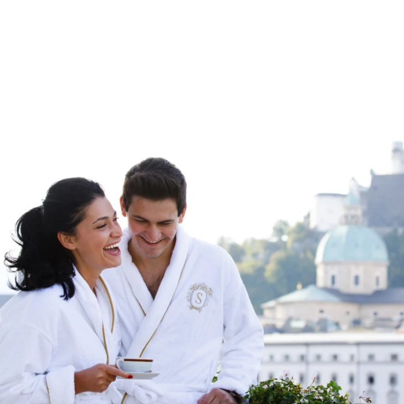 A couple in white bathrobes with the Sacher logo are standing on the balcony. They are both laughing and enjoying a cup of coffee together. In the background, you can see the silhouette of Salzburg with the Hohensalzburg Fortress and the domes of the cathedral.