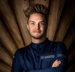 A picture of cook Stefan Fischer with a darf background made out of wood.