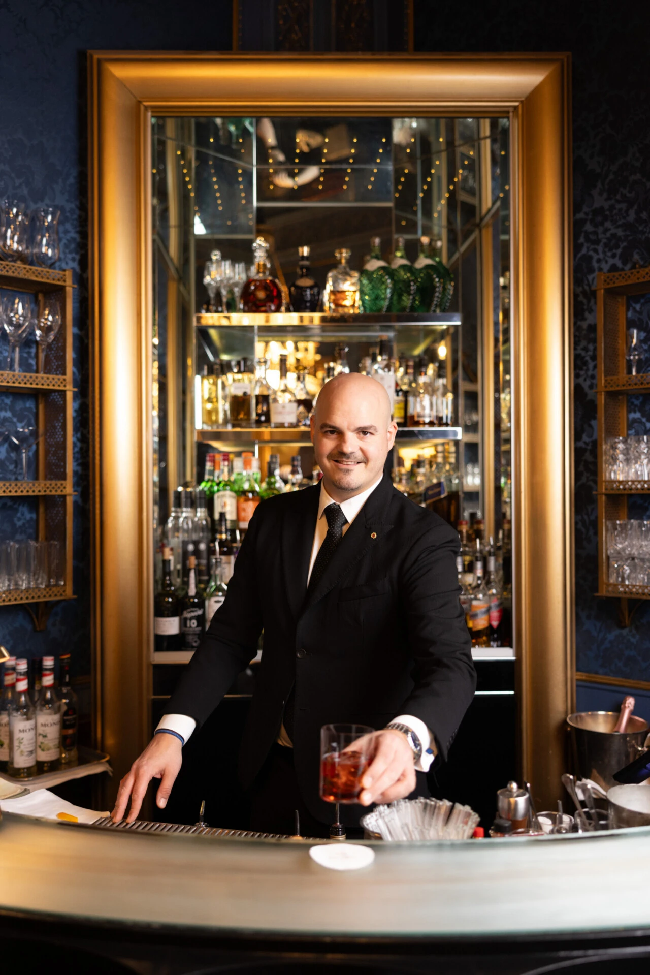 Bartender in a suit serving a cocktail at an elegant bar with a mirrored back and liquor shelves.