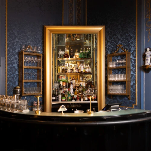 Elegant bar with a curved counter, gold-framed mirror, and well-stocked liquor shelves against a blue patterned wall.