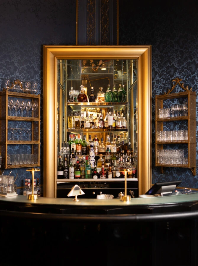 Elegant bar with a curved counter, gold-framed mirror, and well-stocked liquor shelves against a blue patterned wall.