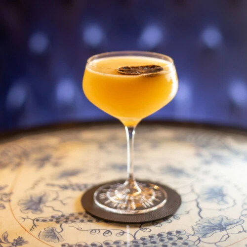 Cocktail in a coupe glass with dried citrus garnish on a coaster on a decorative table.