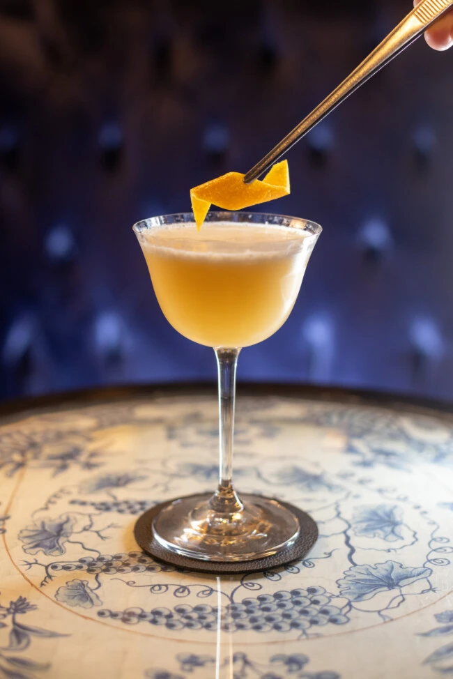 Cocktail in a coupe glass garnished with an orange twist held above the drink with tweezers, on a coaster on a decorative table.