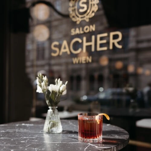 Negroni cocktail on marble table inside Salon Sacher Wien with gold logo on window and Vienna city reflections.