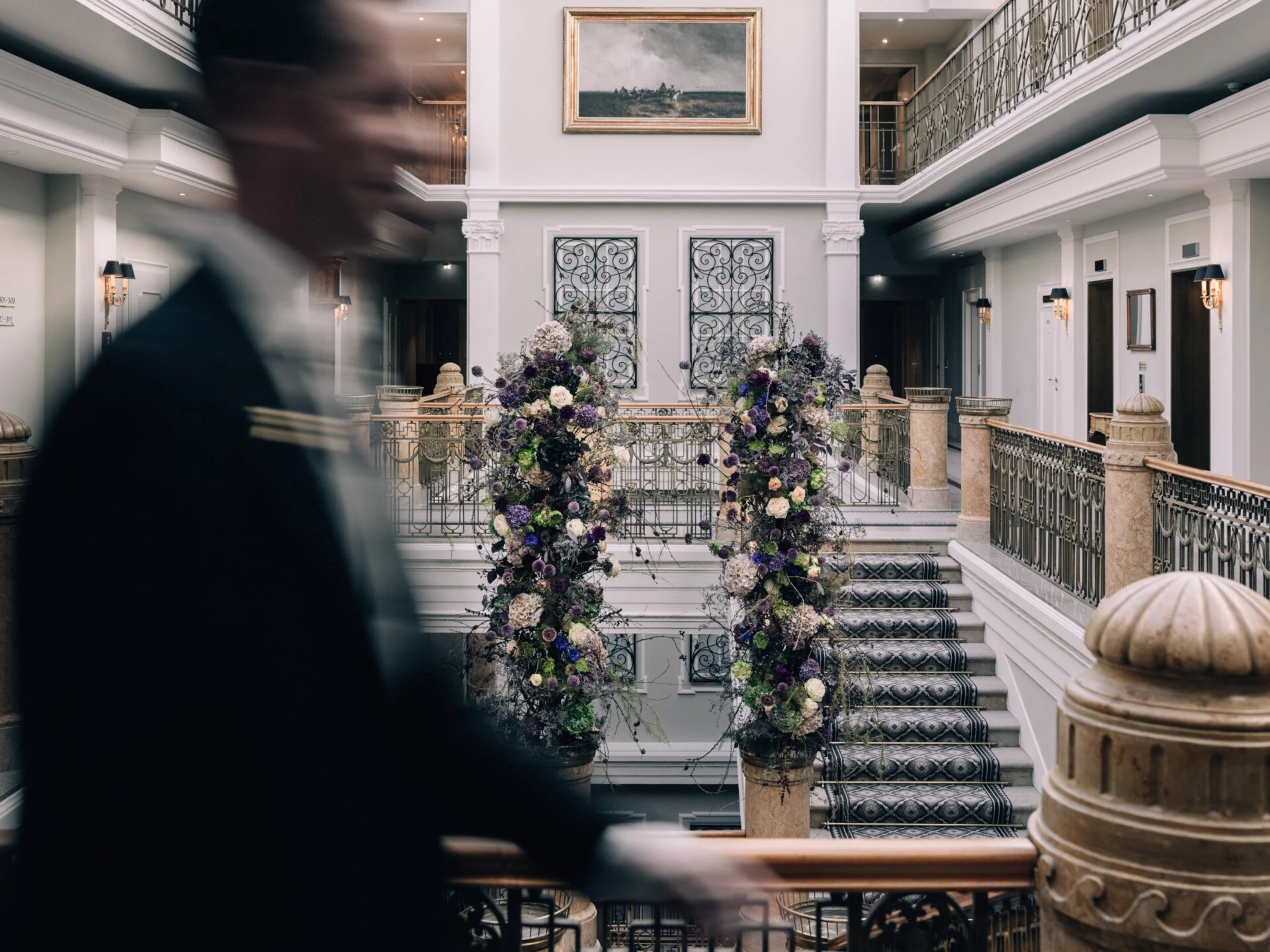 Elegant hotel atrium with a staircase and floral decorations, with a person passing by in motion in the foreground